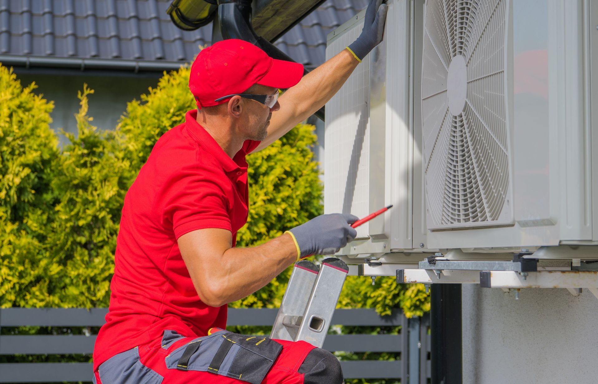 HVAC technician in red shirt and cap, inspecting an outdoor air conditioning unit with a screwdriver.