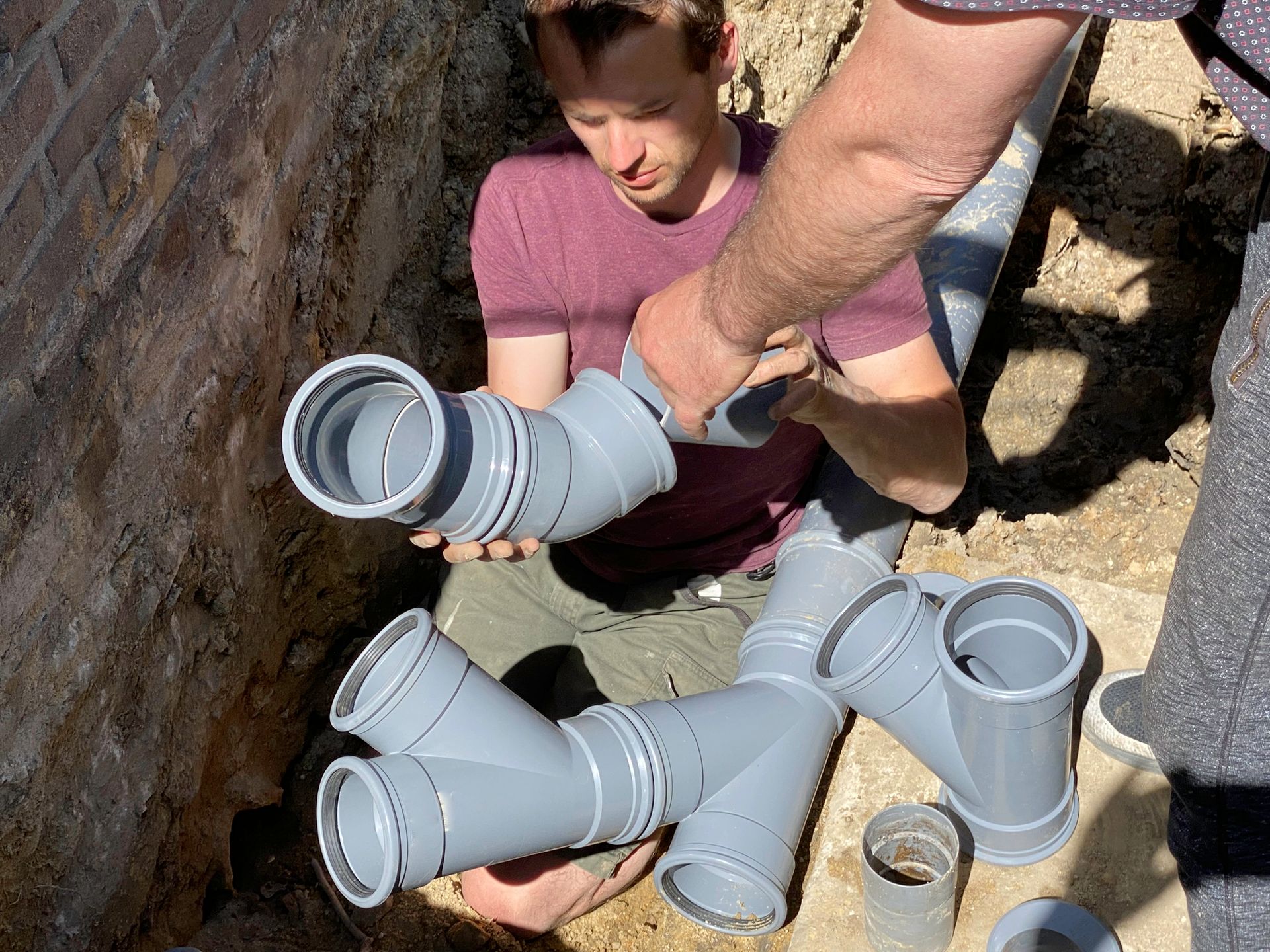 Two people assembling gray PVC pipes in a trench next to a brick wall; construction work.
