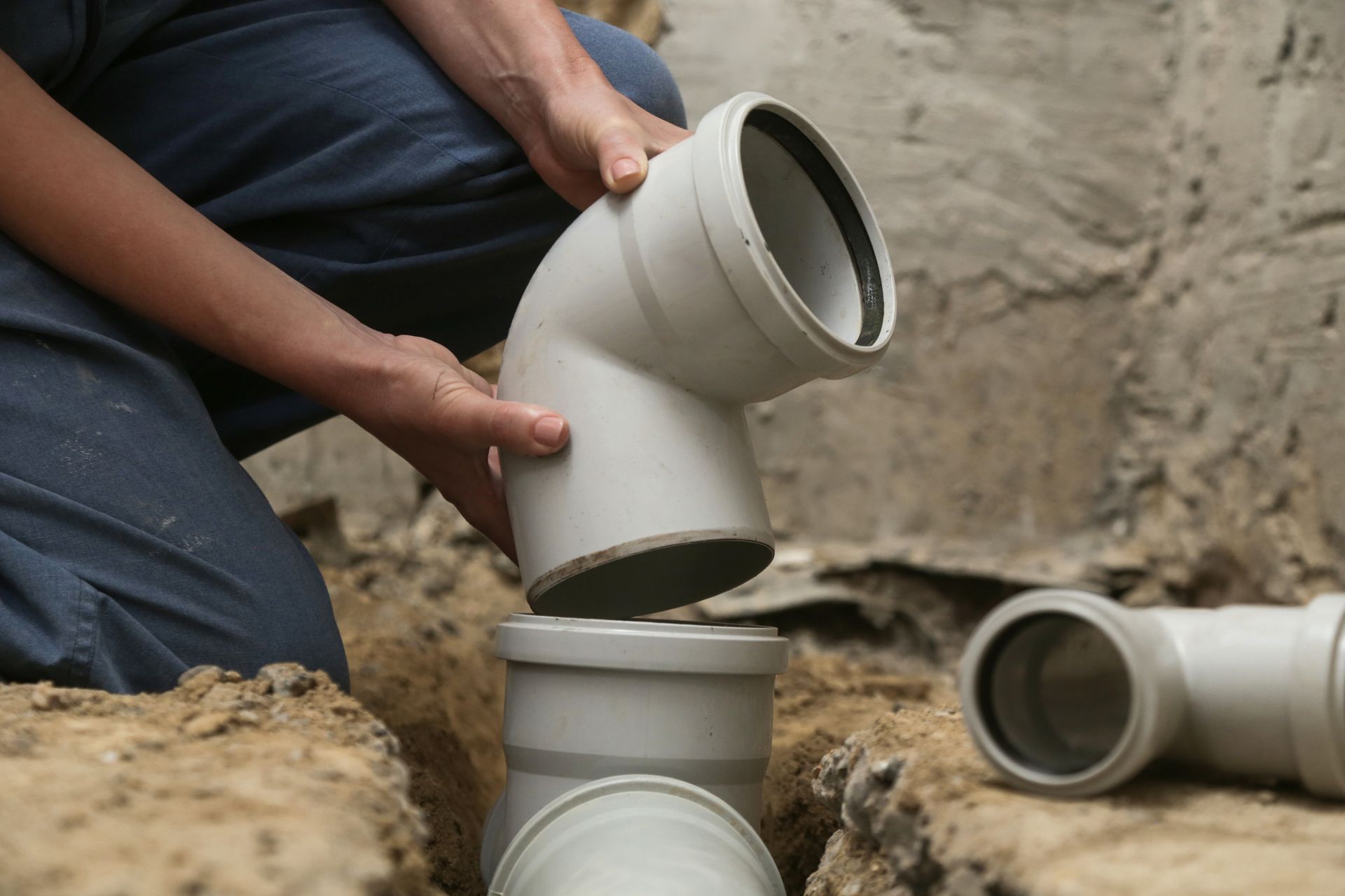 Person connecting gray PVC pipe elbow to a pipe in a trench.
