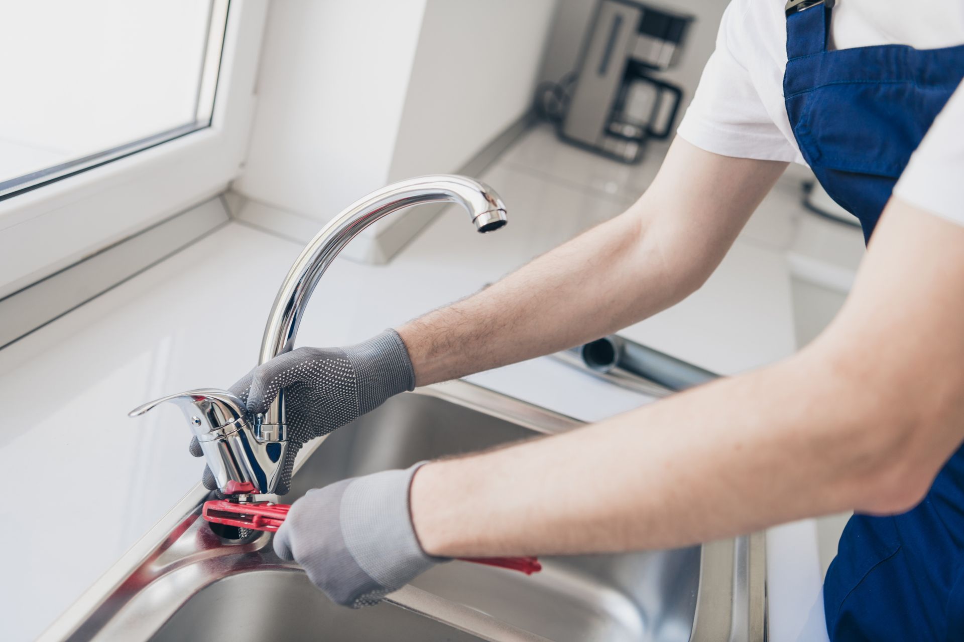 Plumber in blue overalls uses a wrench to repair a faucet in a stainless steel sink.