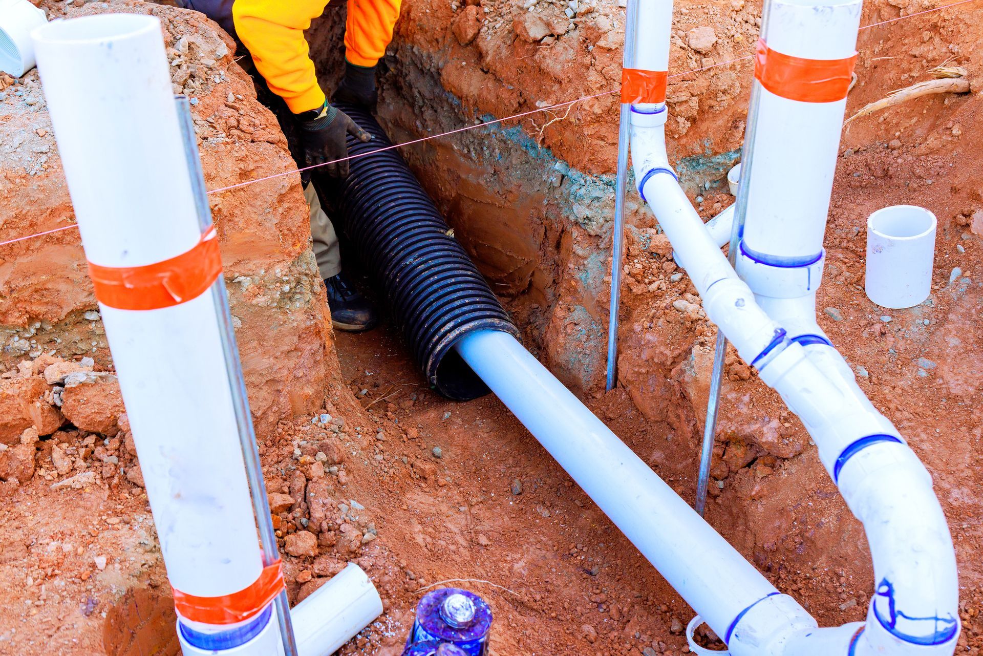 Plumbing installation in progress: white and black pipes in a trench. A worker is also present.