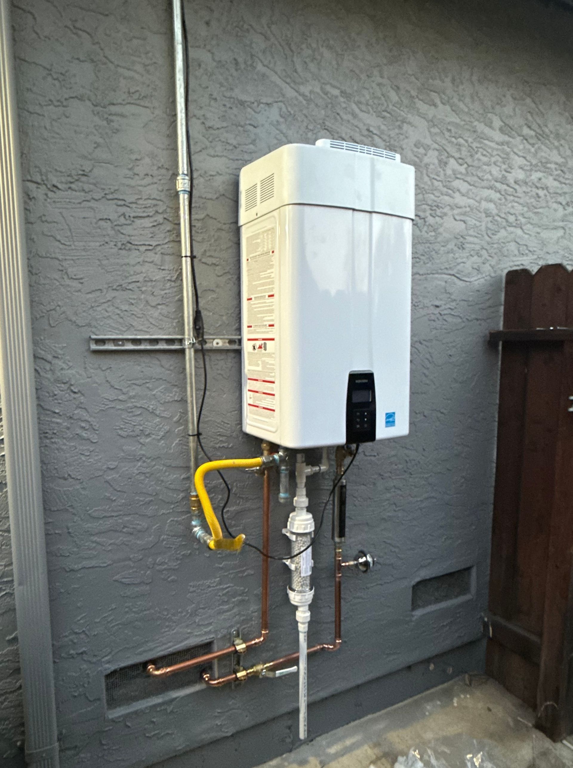 White tankless water heater mounted on a gray exterior wall, with copper and yellow gas pipes.