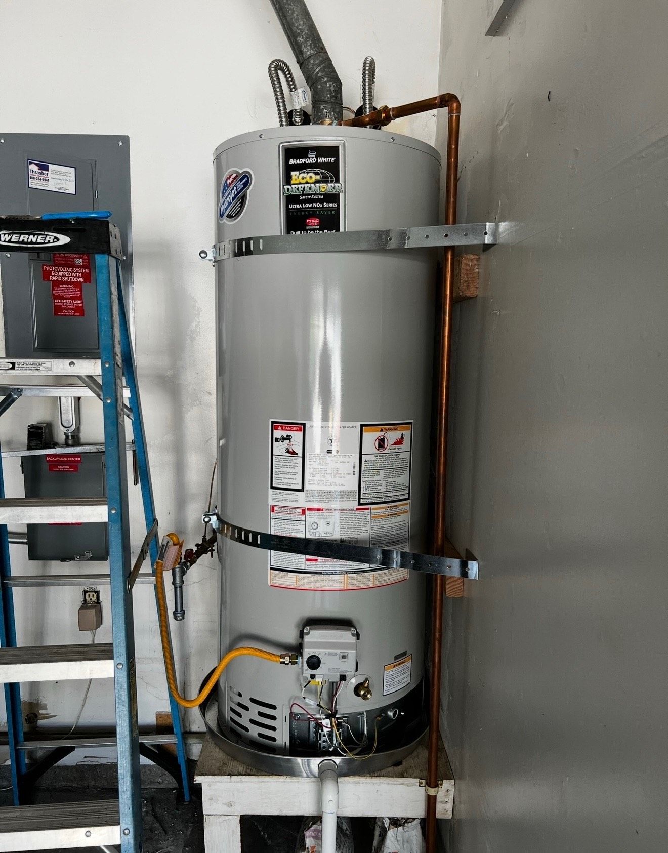 Tall gray water heater in a garage, secured to the wall with metal straps. Copper pipes run alongside. A ladder stands next to it.