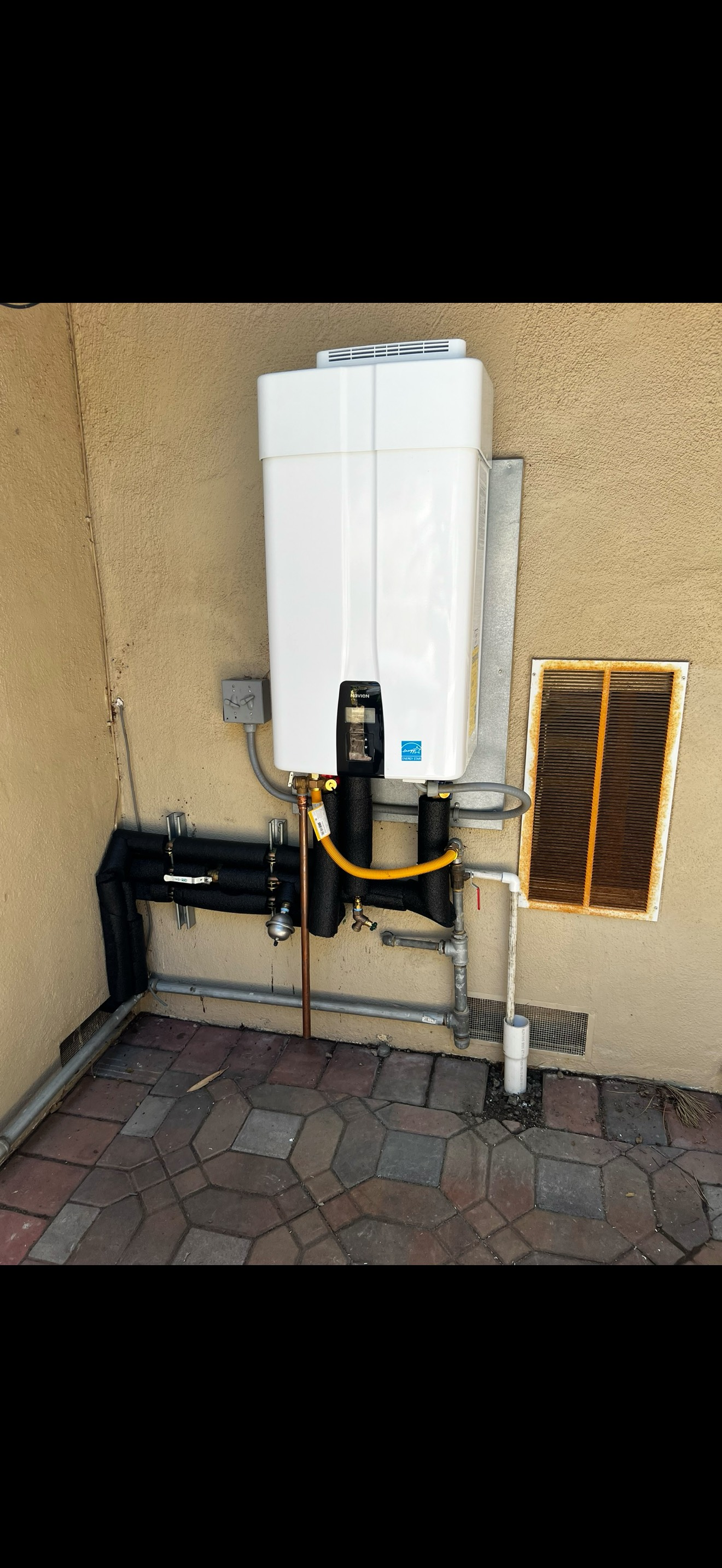 A white water heater attached to a tan wall, with black pipes, a yellow hose, and a vent.