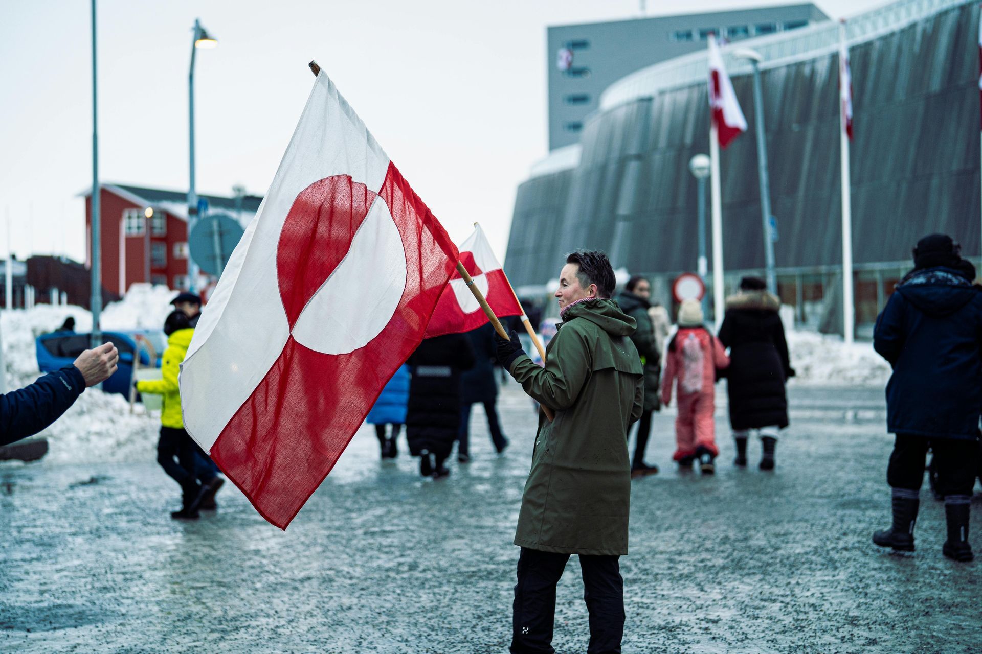 Man waving Greenlandic flag in front of a building; other people in the background. White and red flag.