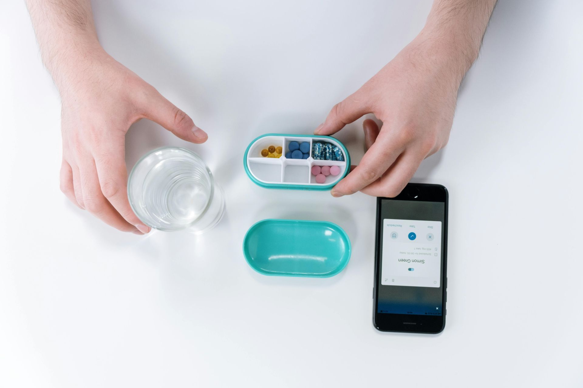 Hands near pill organizer, glass of water, and smartphone on white surface. The phone shows a reminder screen.