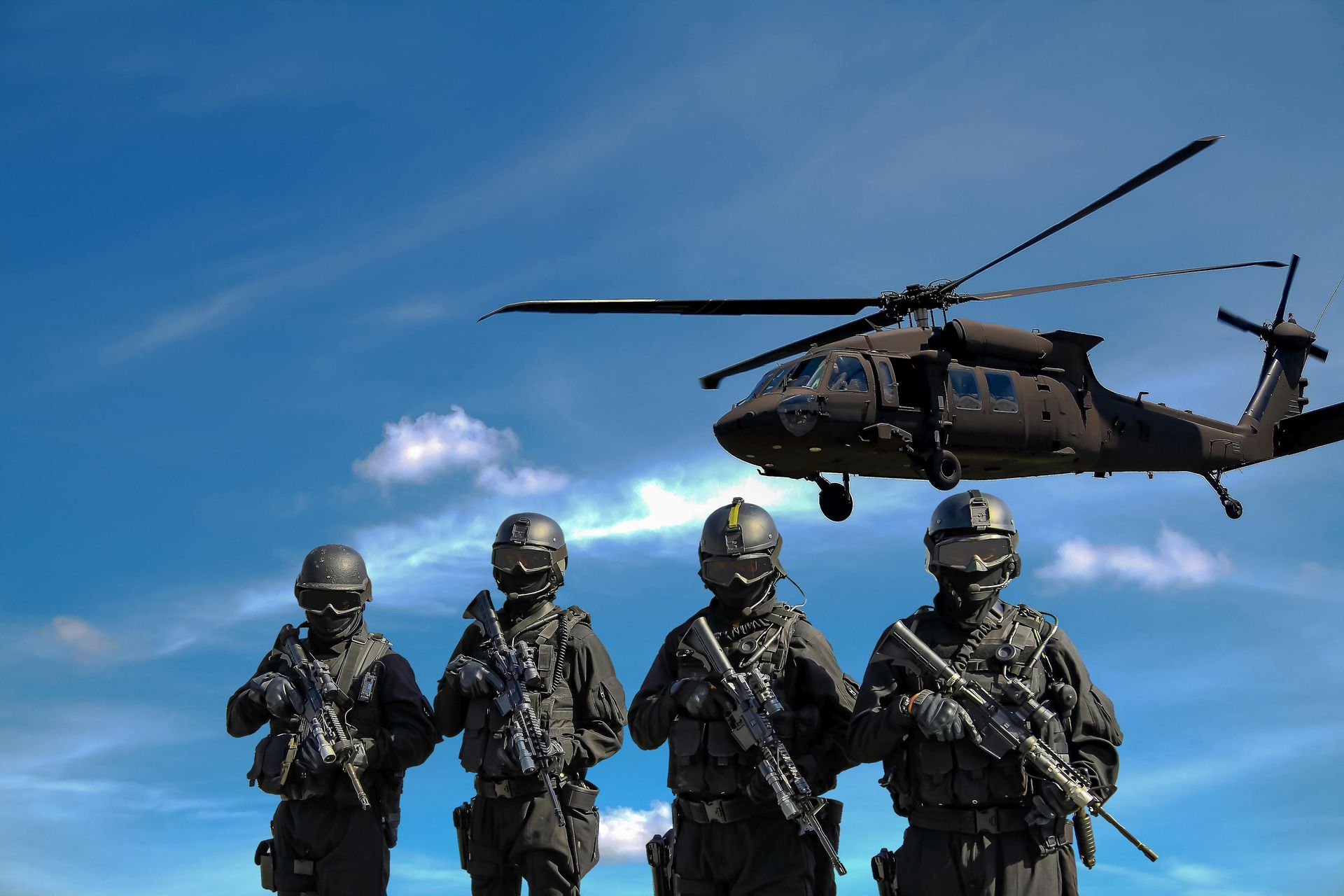 Four armed soldiers in tactical gear stand beneath a hovering black helicopter against a blue sky.