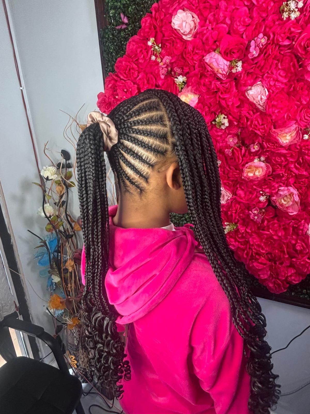 A person wearing a high ponytail with braided hair and loose, curly ends, standing in front of a bright pink rose wall.