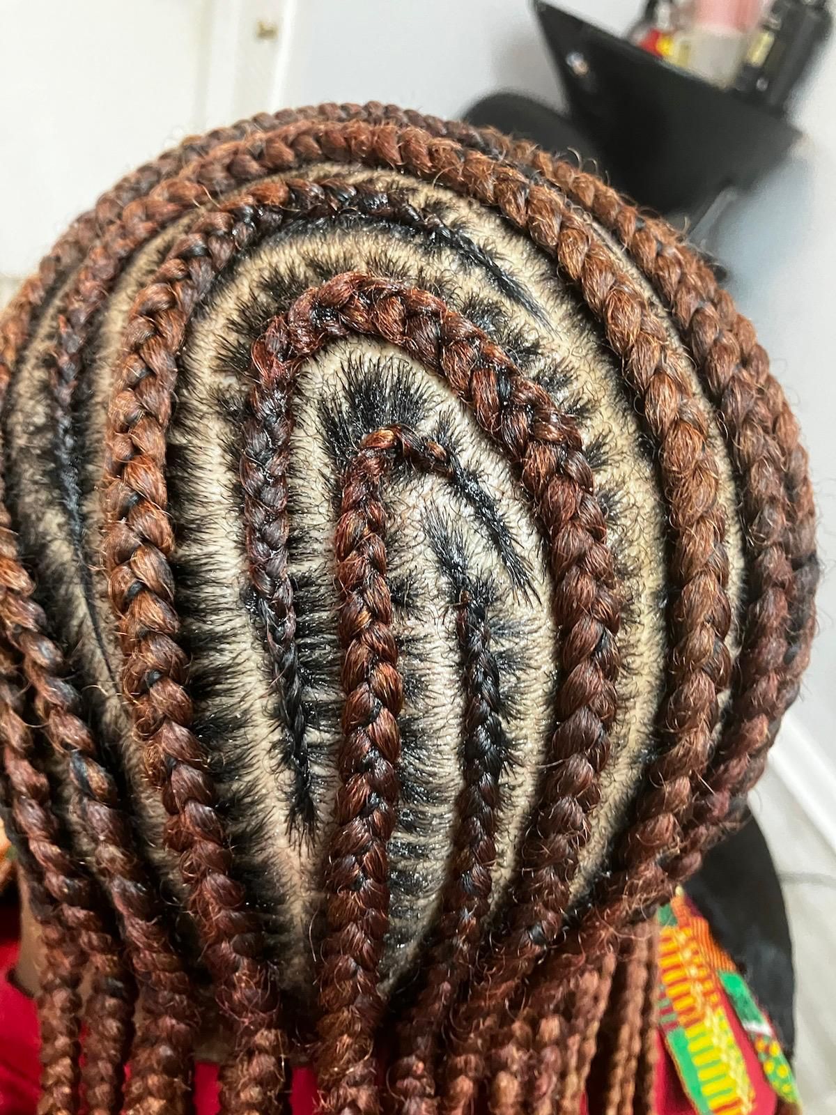 A close-up of hair styled in intricate cornrows that curve in a spiral pattern, colored in shades of brown.