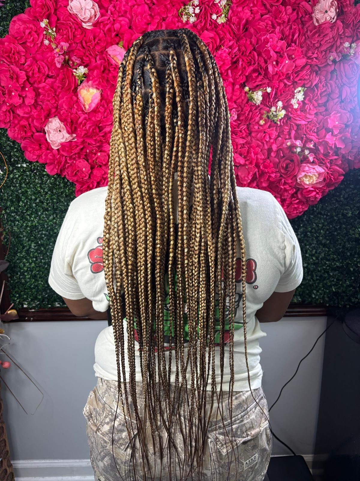 Back view of long, honey-blonde knotless braids against a backdrop of artificial red flowers and green foliage.