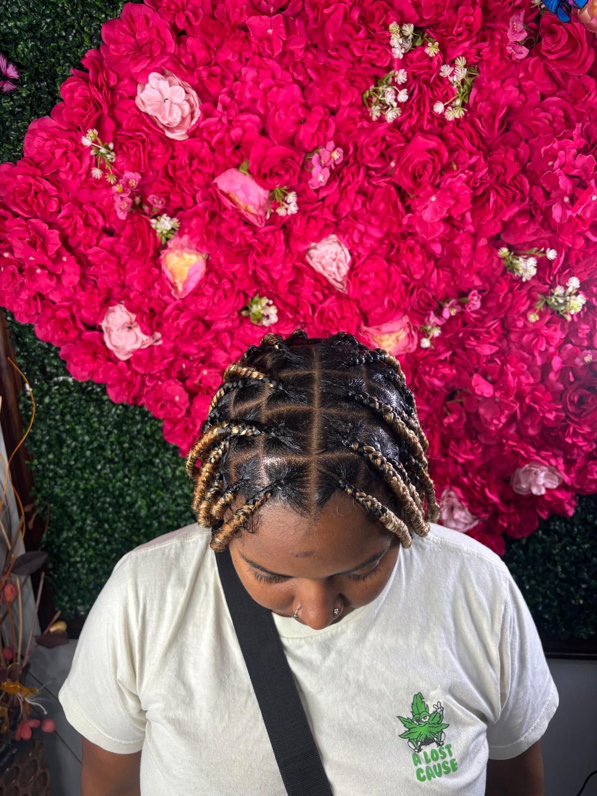 A person with blonde-tipped braided hair styled in neat, square sections against a wall of vibrant red roses.
