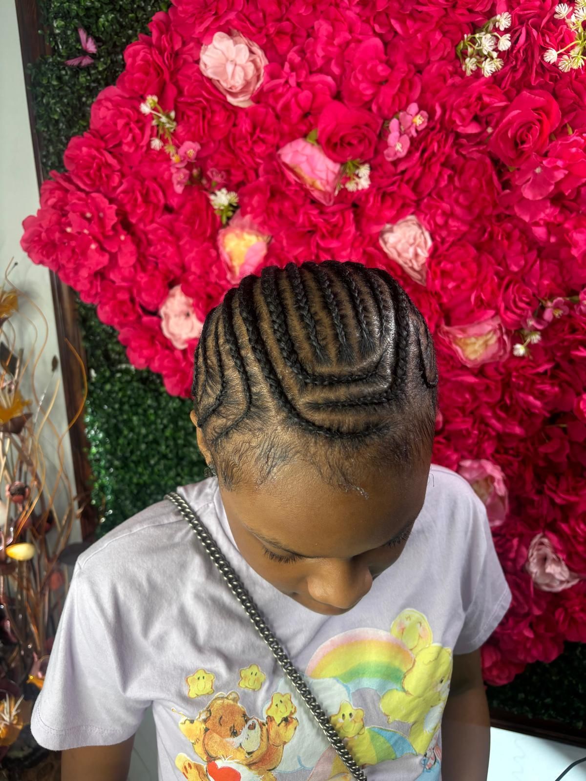 A person with intricate, curved braided hair standing in front of a wall of vibrant red and pink faux flowers.