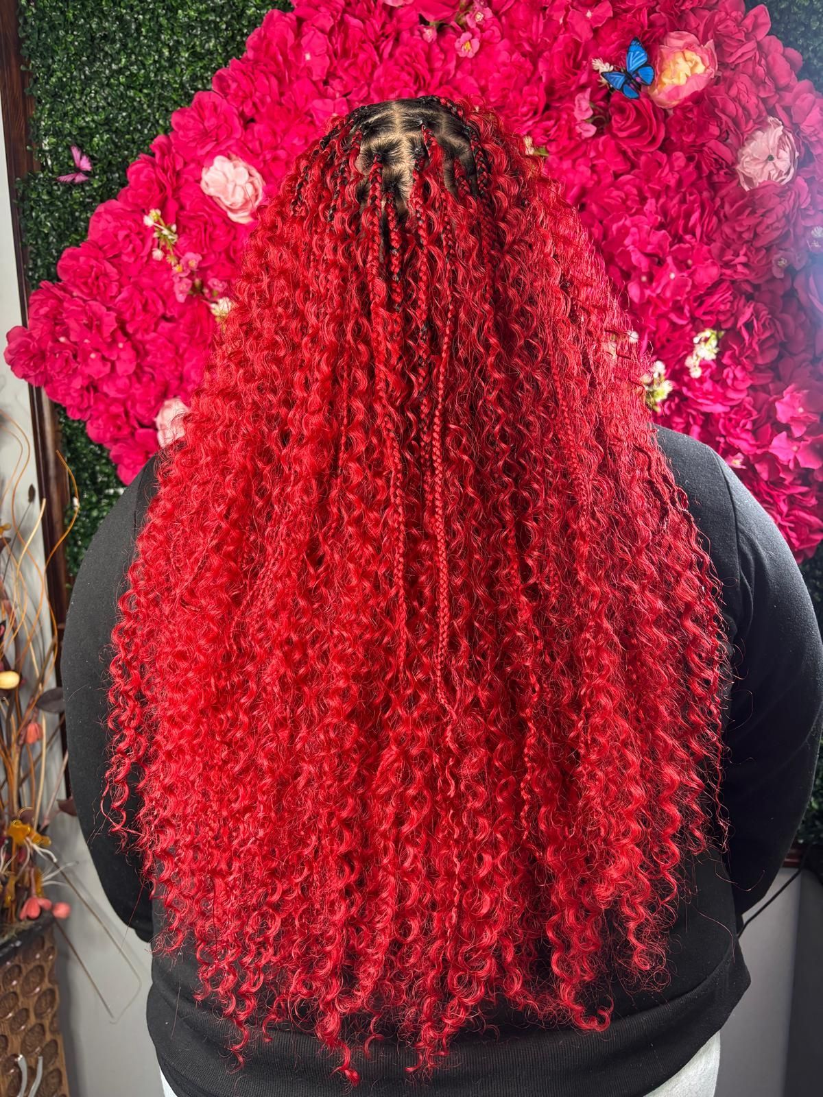 A person with long, vibrant red curly box braids standing in front of a backdrop of red flowers.