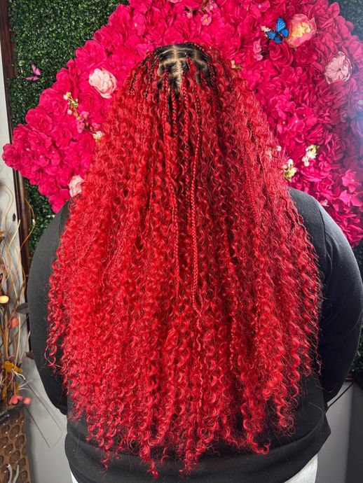 A person with long, vibrant red curly hair styled in box braids against a bright red floral background.