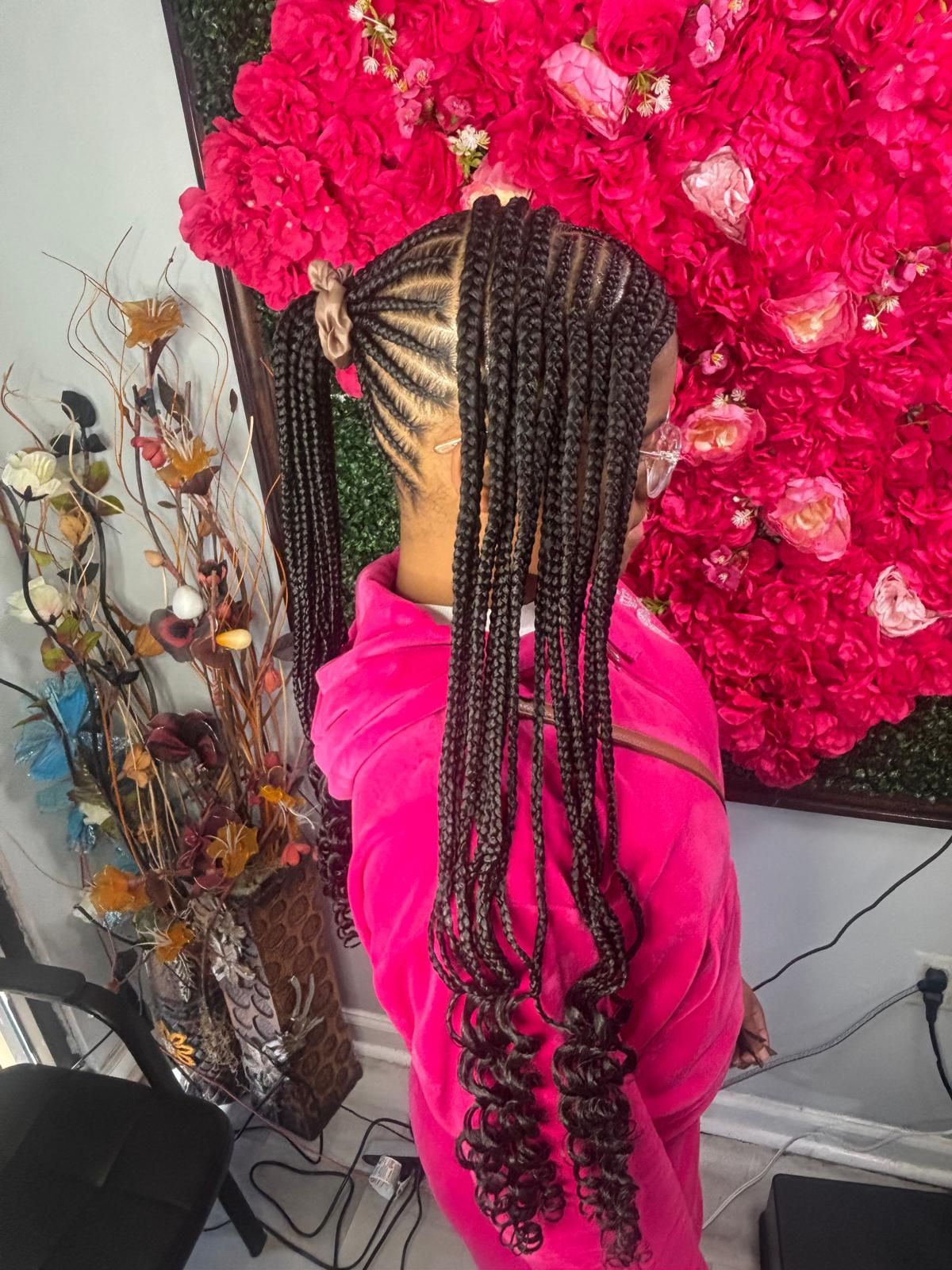 A person with long, dark, braided hair styled into two high ponytails with curly ends, posing against a red floral wall.