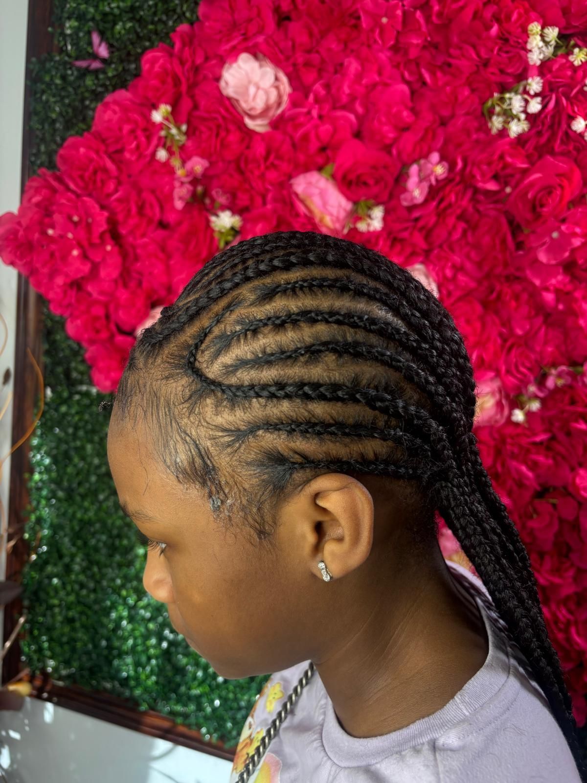 Side profile of hair styled in neat, dark braided cornrows against a backdrop of vibrant pink artificial roses.