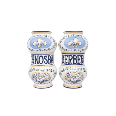 Twee keramische apothekerspotten in crème en blauw, versierd met leliemotieven, zwanenafbeeldingen en botanische details.