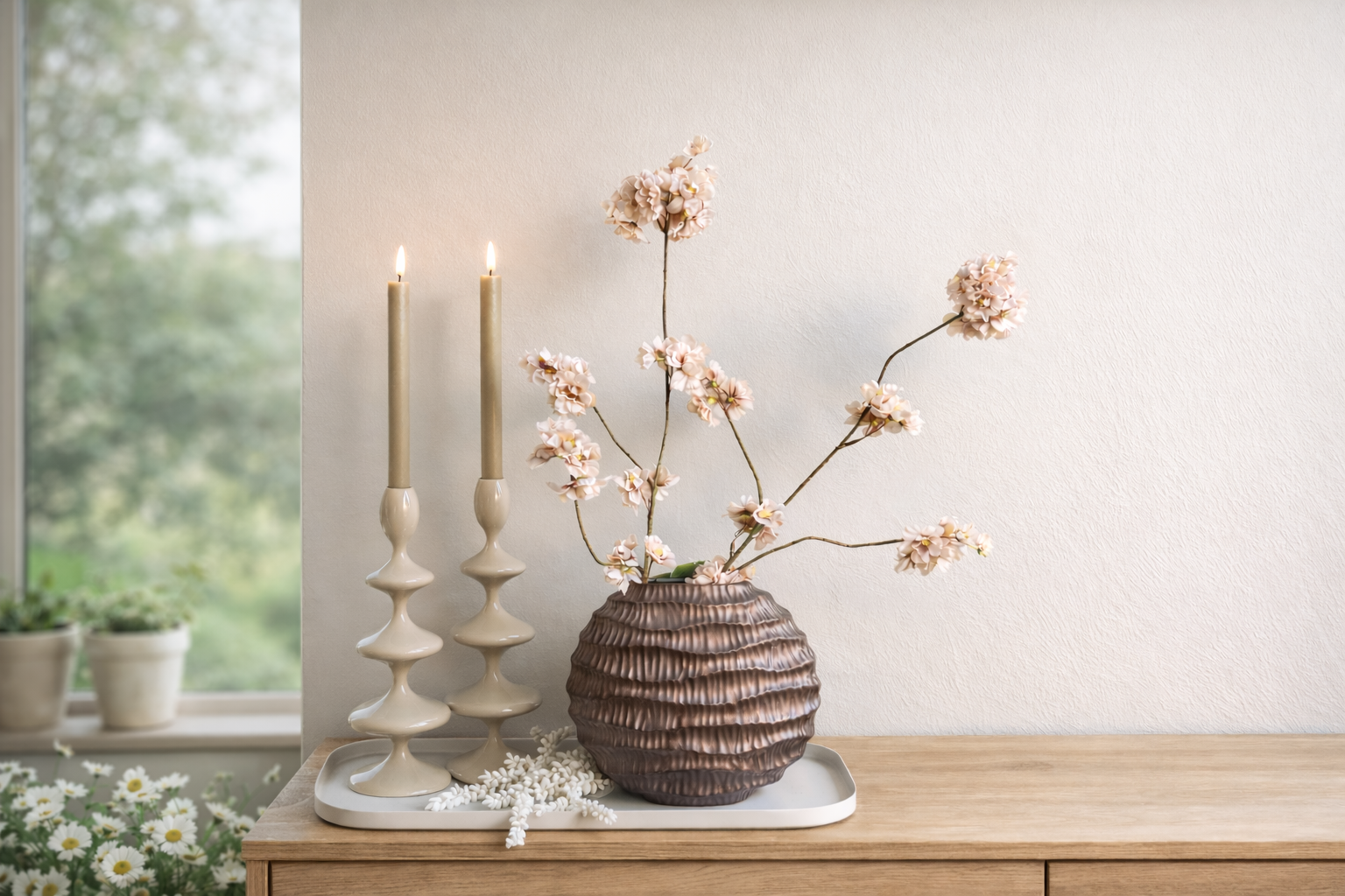 Twee beige kandelaars met brandende kaarsen naast een getextureerde bruine vaas met delicate bloemtakjes op een wit dienblad.
