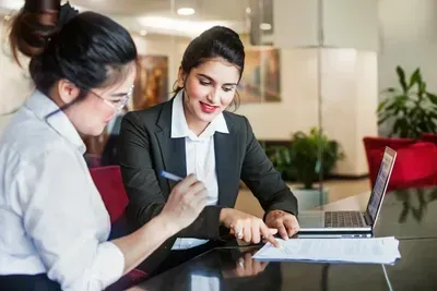 Two women are sitting at a table looking at a laptop computer.