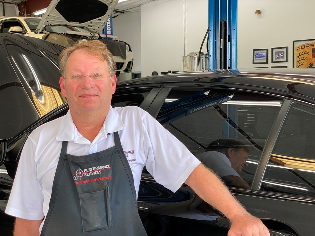 Mechanic standing beside a black car in an auto repair shop, wearing a white shirt and apron