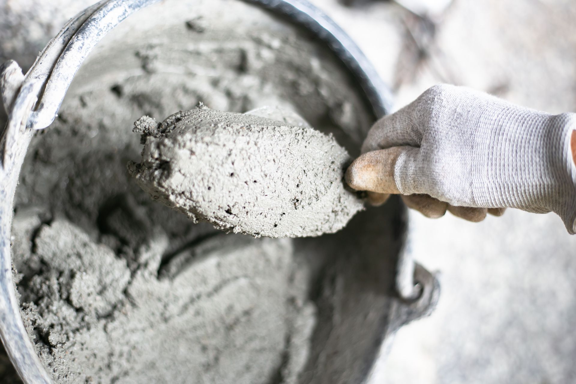 Gloved hand holding a trowel with cement, inside a gray bucket.