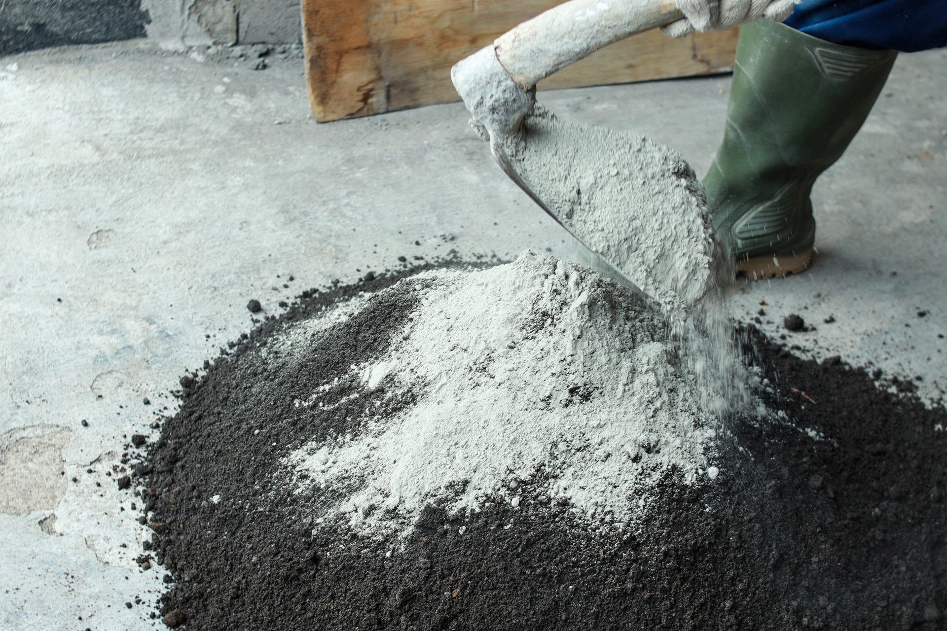 Person mixing cement on the ground with a hoe; gray cement and black soil visible.