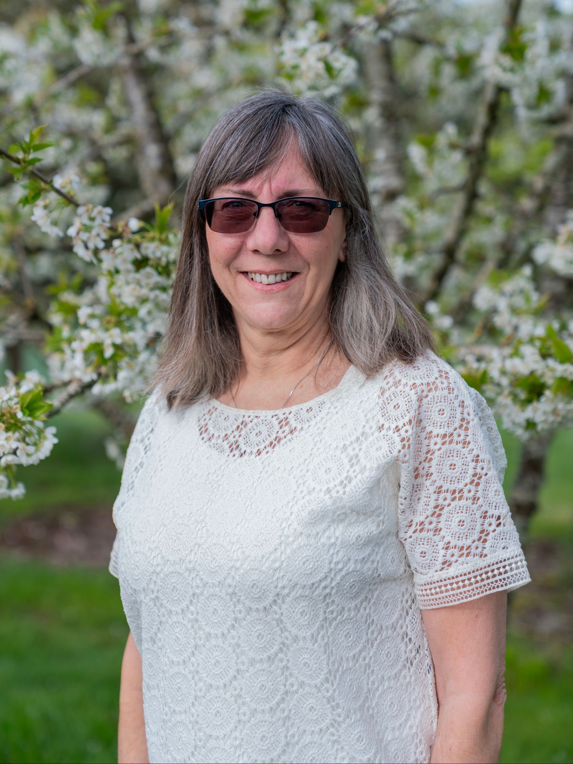 A woman wearing sunglasses and a white lace top is standing in front of a flowering tree.