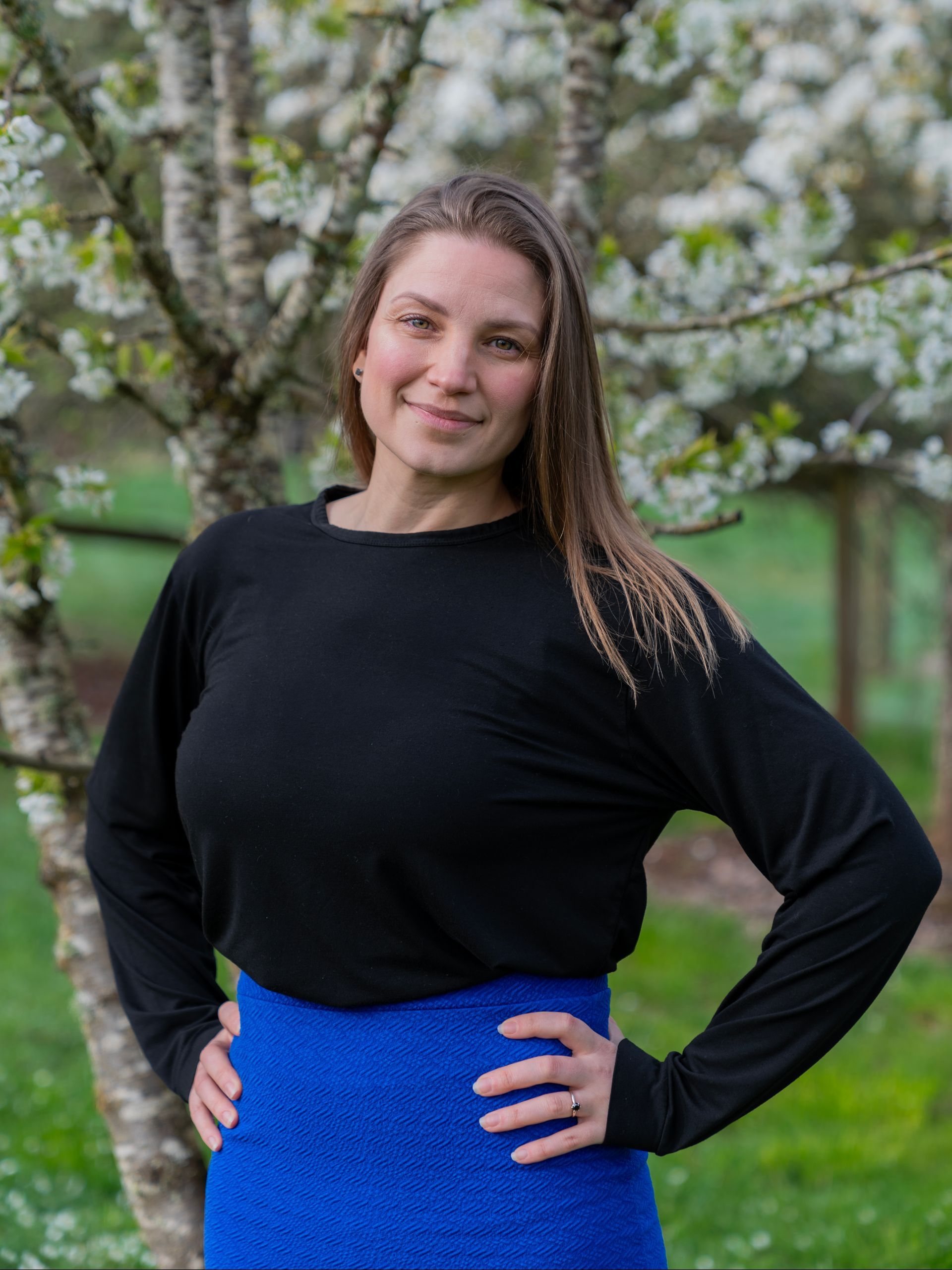 A woman in a black shirt and blue skirt is standing in front of a tree.