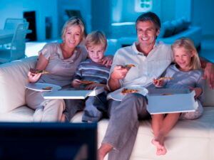 A family is sitting on a couch eating pizza and watching tv.