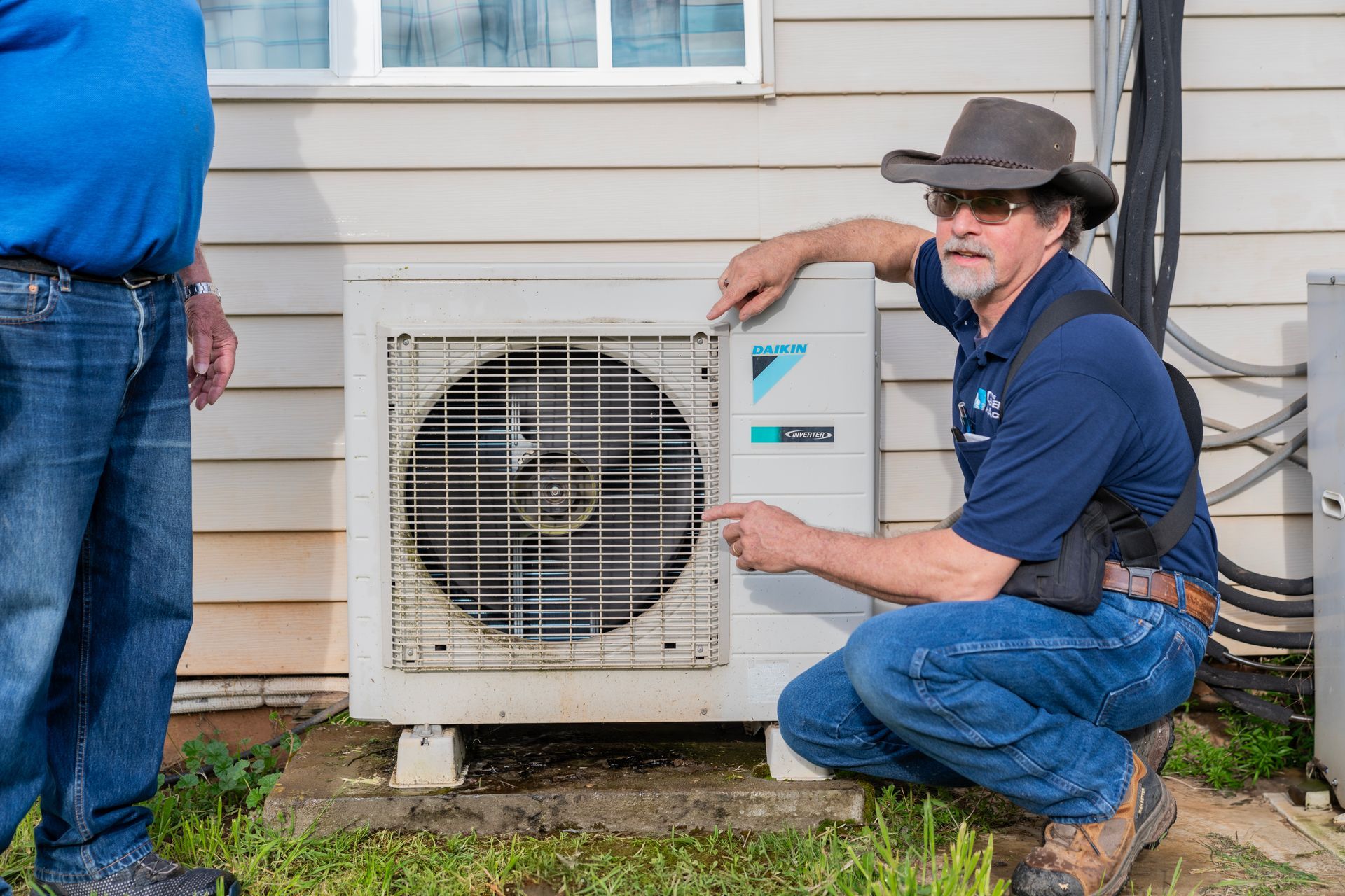 The Clean Air Act Technician in a cowboy hat is working on an air conditioner outside of a house.
