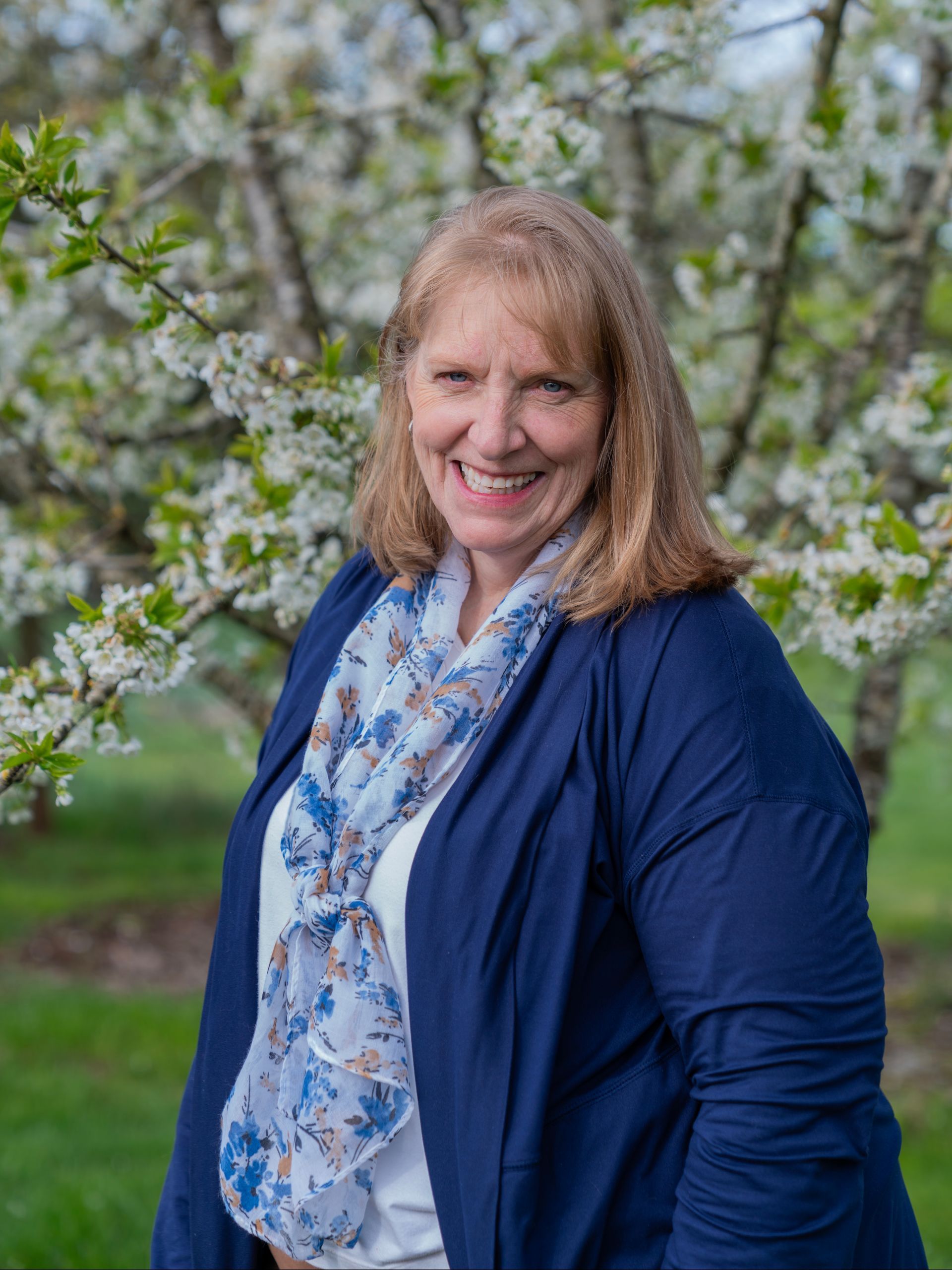A woman in a blue jacket and scarf is standing in front of a tree with white flowers.