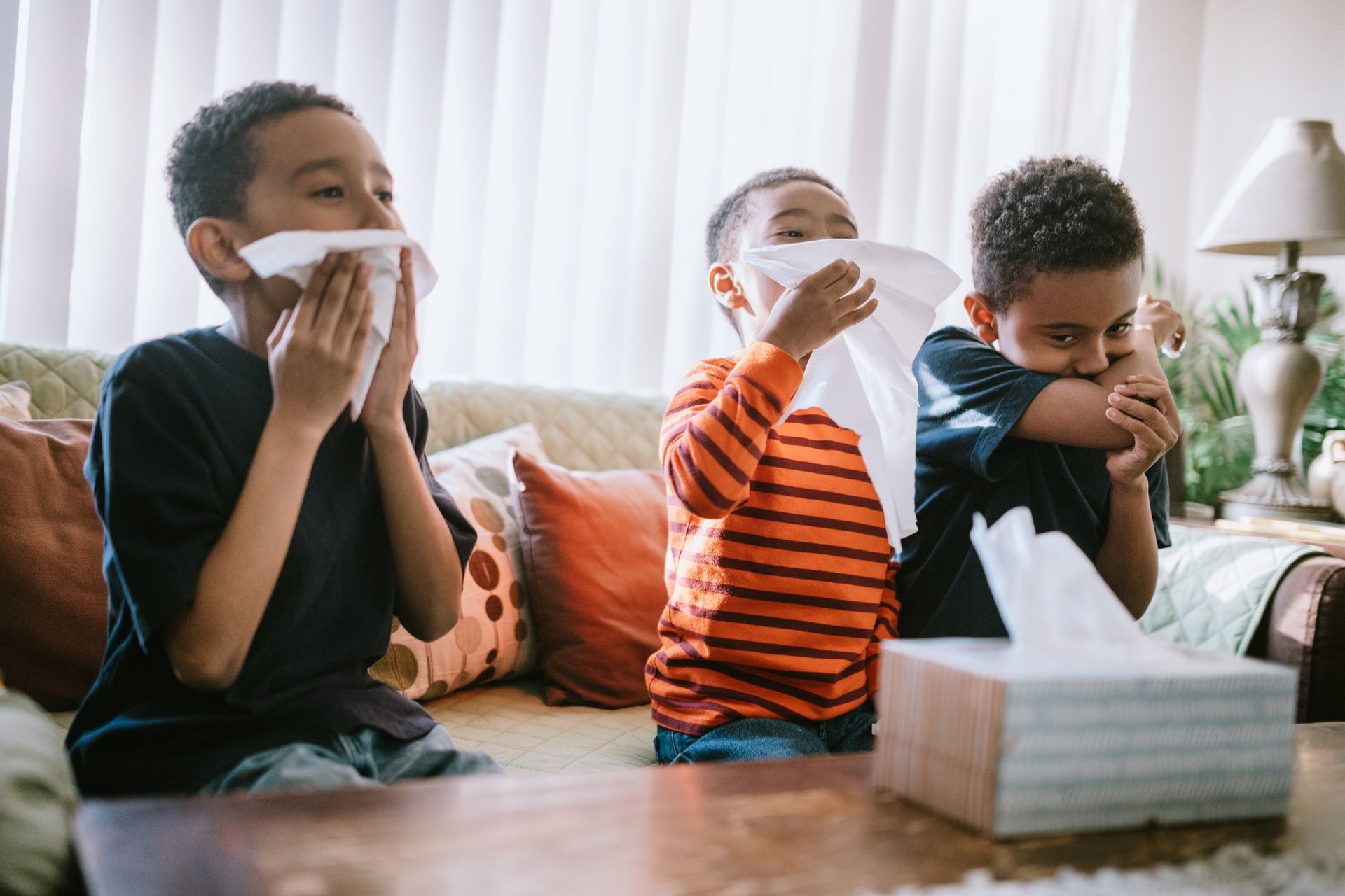 Three young boys are blowing their noses with tissues while sitting on a couch.