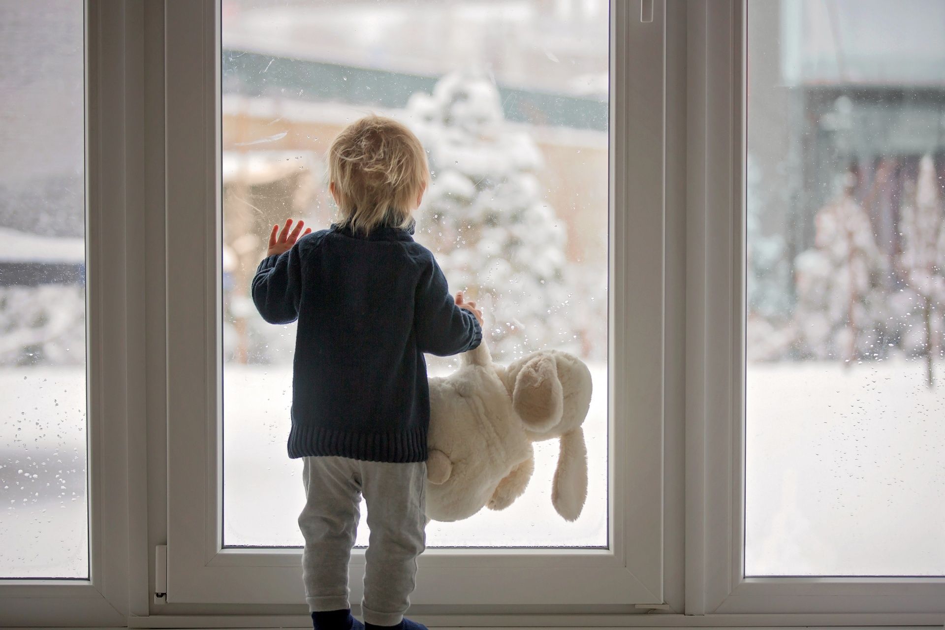 A little boy is holding a teddy bear and looking out of a window at the snow.