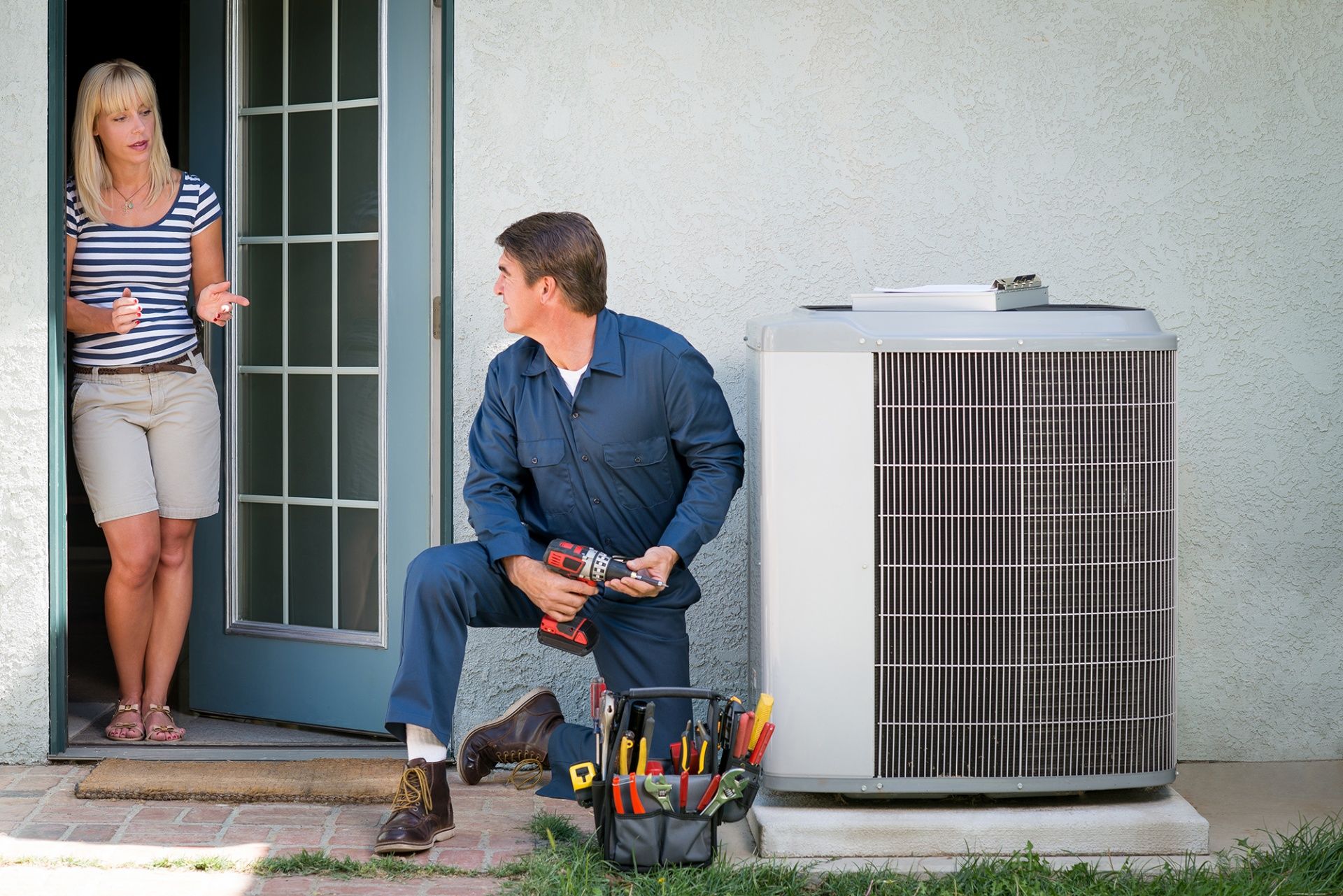 The Clean Air Act Technician is kneeling down next to an air conditioner while a woman stands in the doorway.