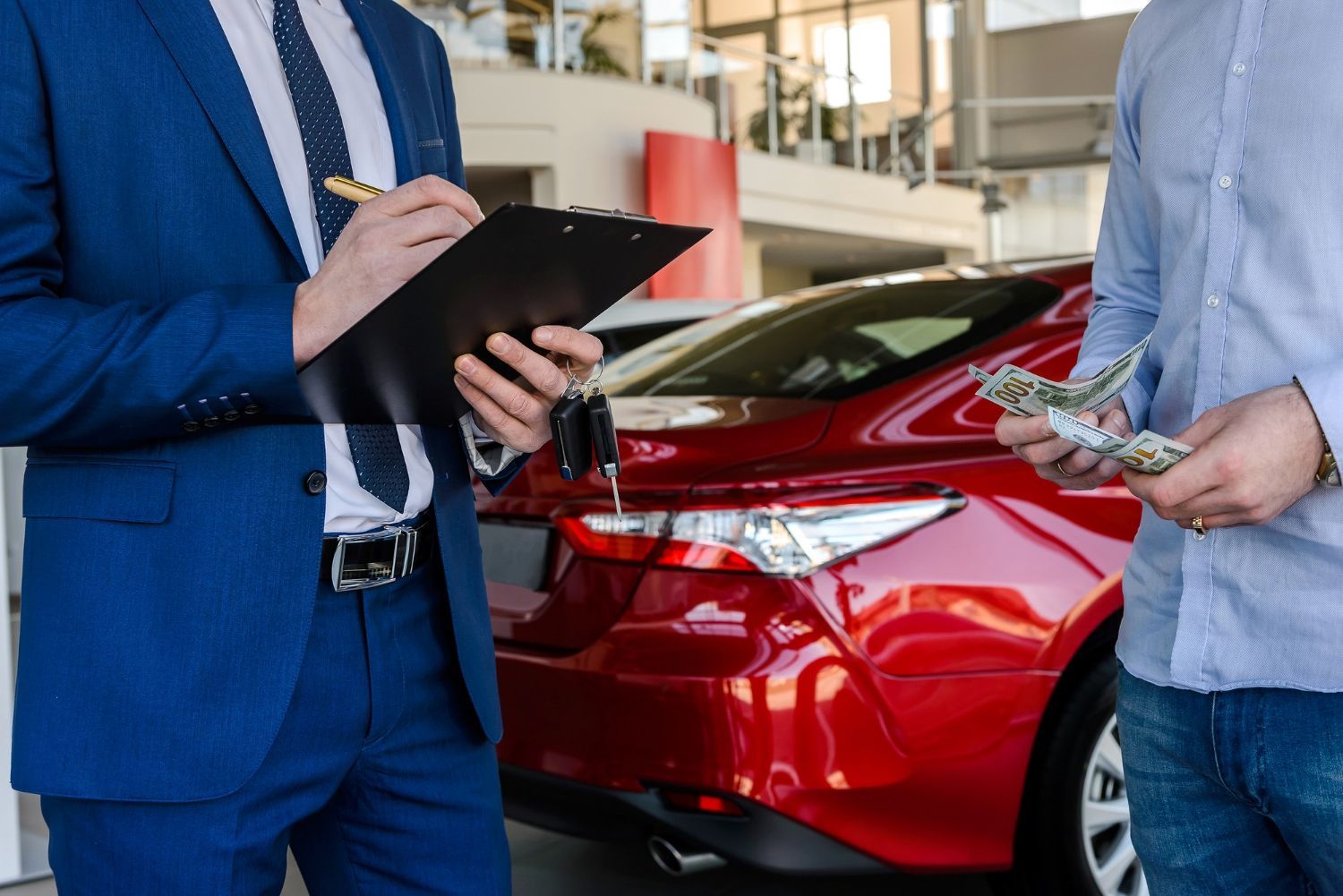 A man in a suit is holding a clipboard and talking to another man in front of a red car.