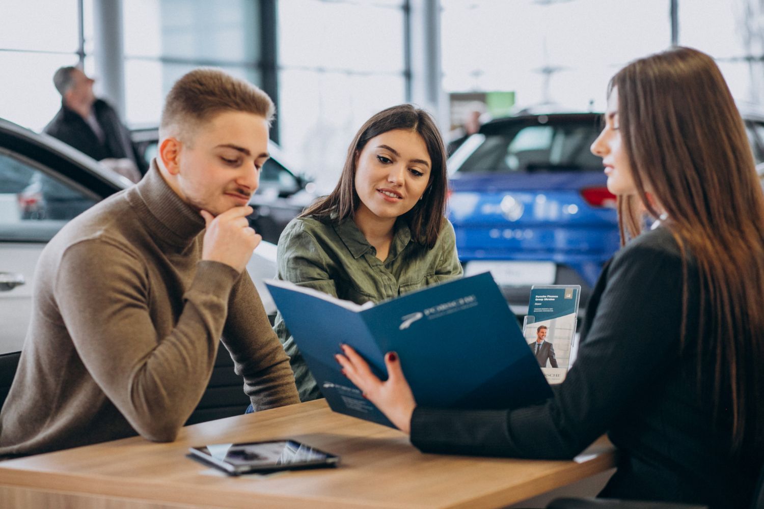 A man and a woman are sitting at a table in a car showroom looking at a book.