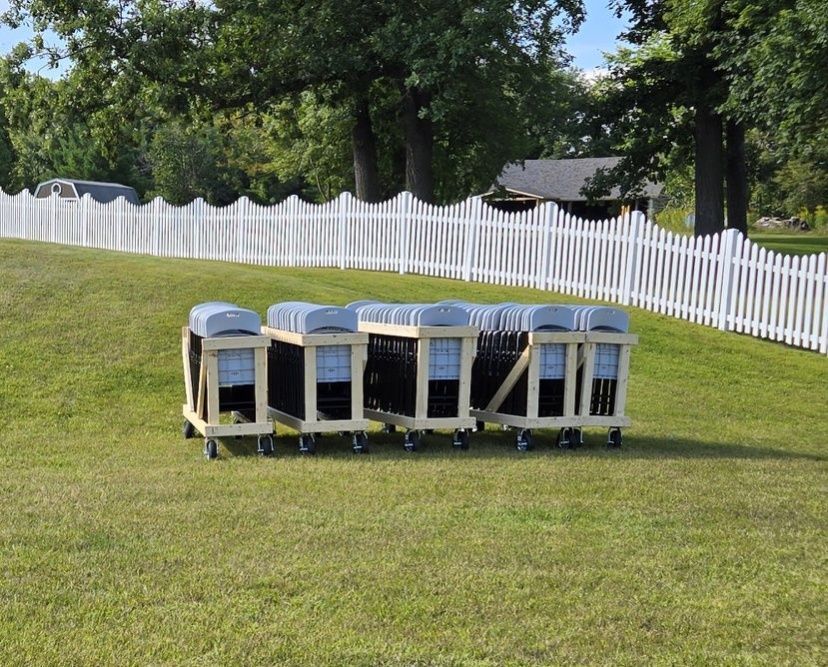 Carts of black folding chairs on a grassy hill, with a white picket fence in the background.