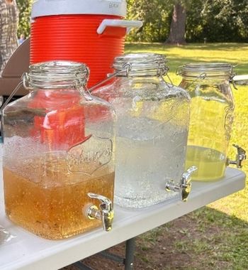 Beverage dispensers with spouts filled with drinks on a white table, cooler in background, outdoor setting.