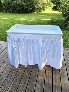 White plastic table with ruffled skirt on a wooden deck in front of greenery.