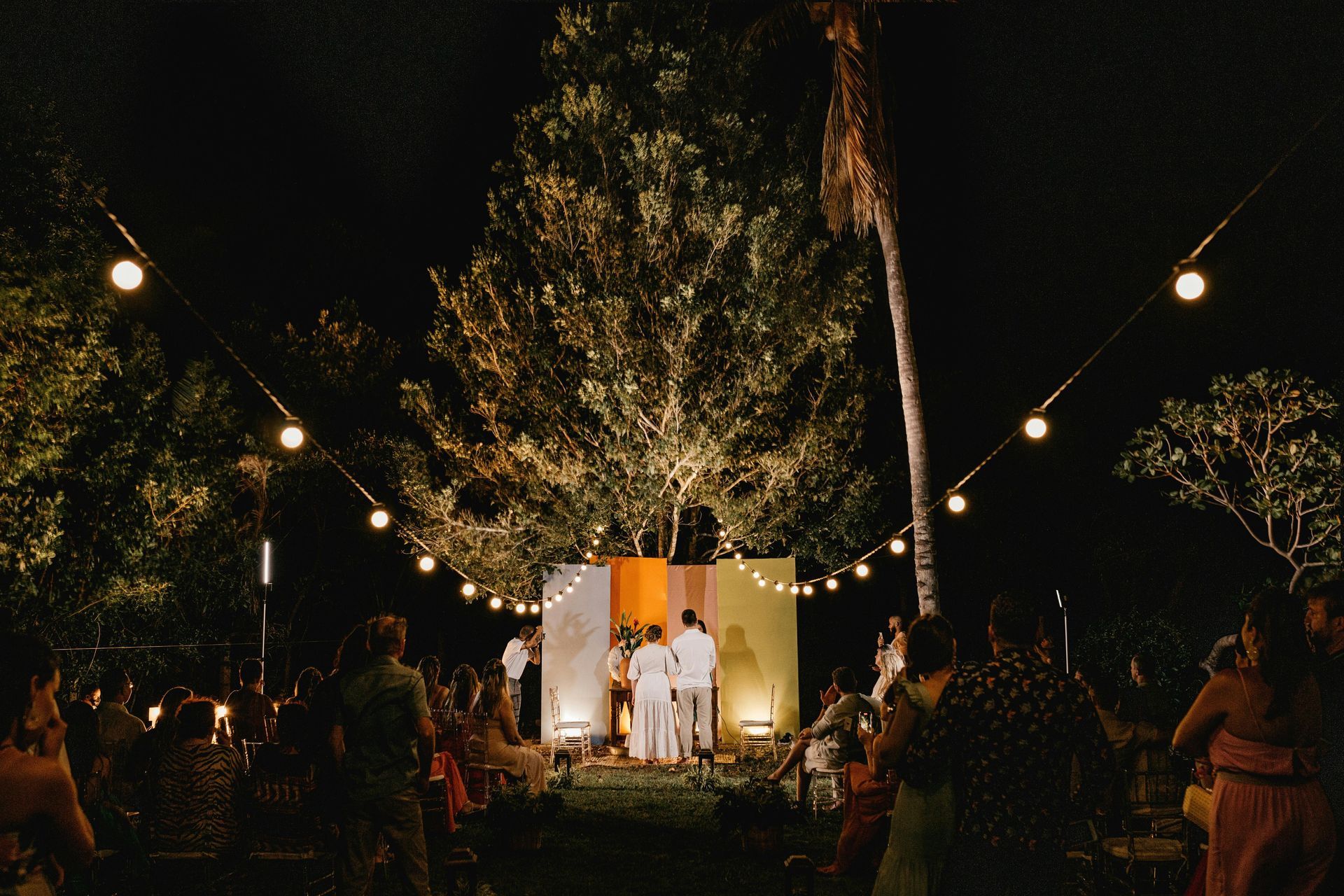 Nighttime wedding ceremony. Guests watch couple stand before colorful backdrop, illuminated by string lights and trees.