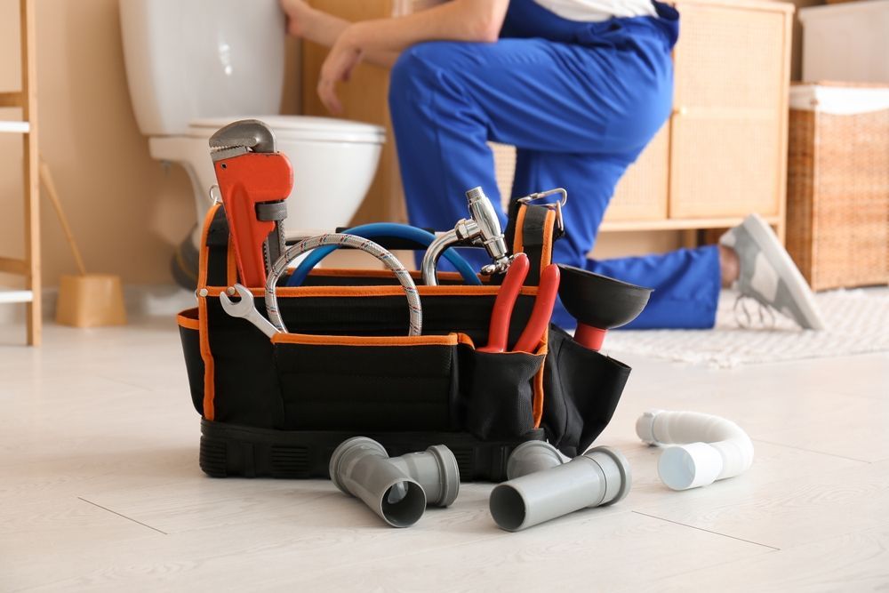 A plumber in blue workwear kneels by a toilet near a tool bag filled with equipment and loose pipe fittings on the floor.