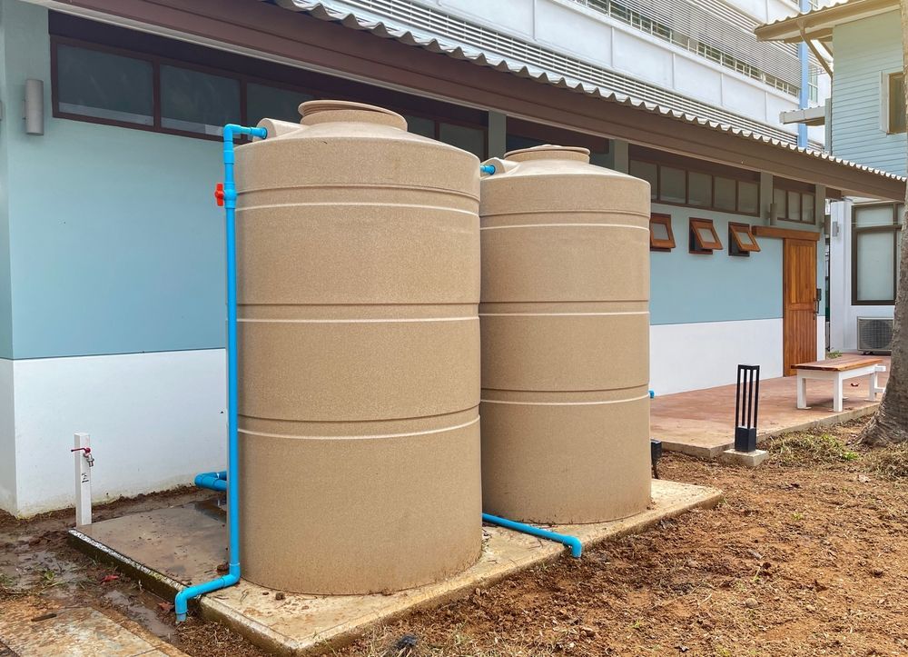 Two large, beige cylindrical water storage tanks sit on a concrete pad outside a light blue building.