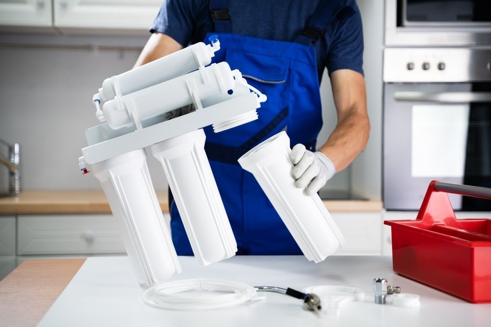 A technician in blue workwear installs a multi-stage water filtration system on a white kitchen countertop.