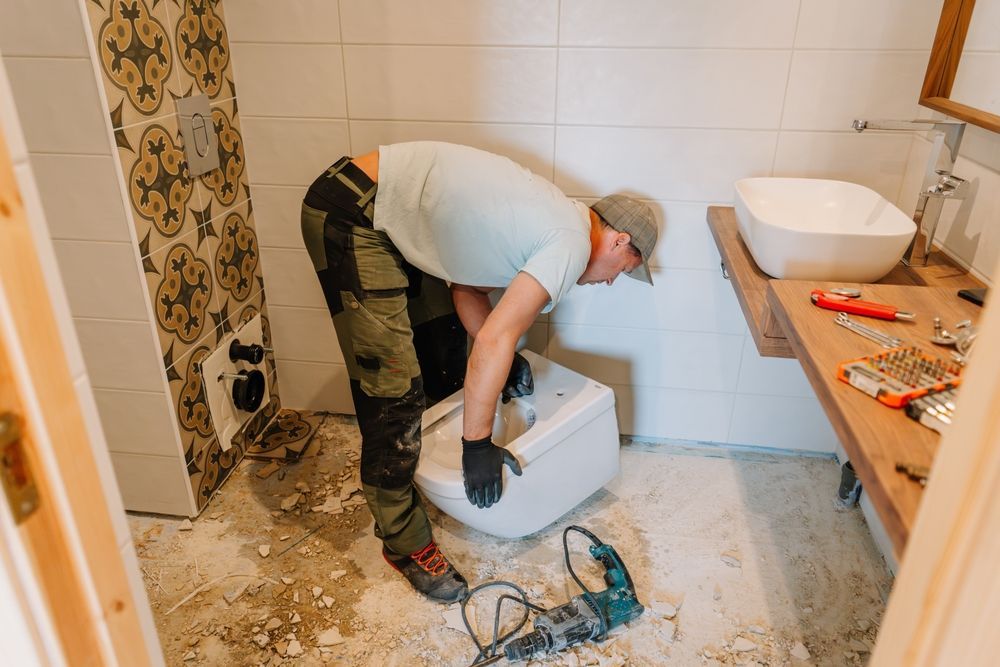 A worker in work clothes installs a wall-mounted toilet in a bathroom under renovation with tiled walls and a sink.
