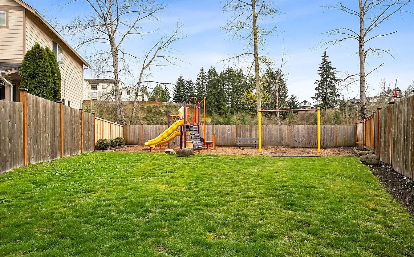 Backyard with a wooden fence, green grass, and a playground.  Houses and trees in the background.