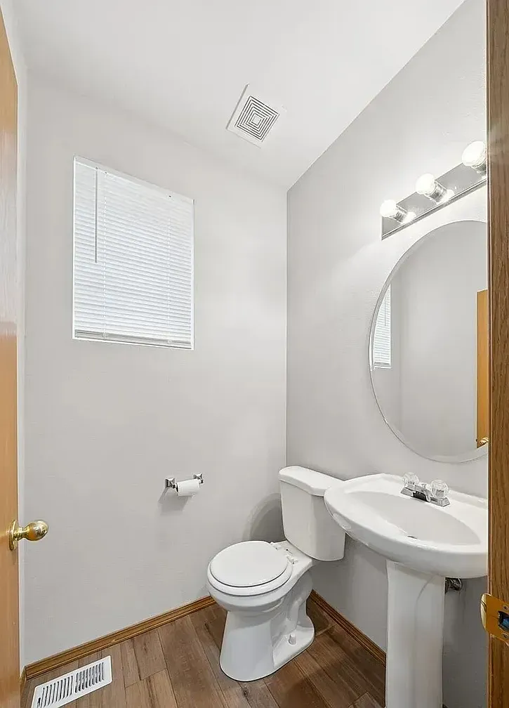 Small bathroom with toilet, pedestal sink, and window. Gray walls, wood-look floor, and a mirror.