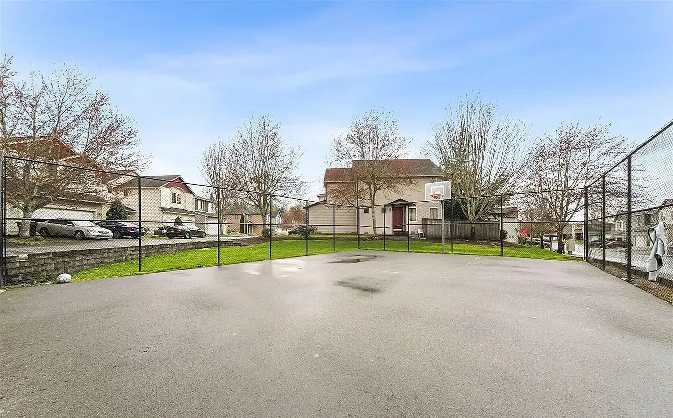 Paved lot with grass and trees. Houses and a fence line the background under a blue sky.