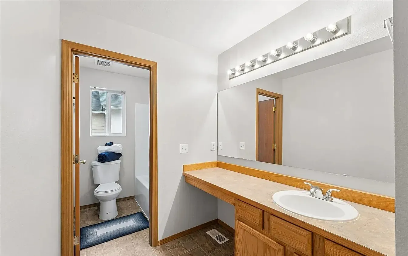 Bathroom with a toilet, vanity with sink, and mirror. White walls, wood trim, and a window.