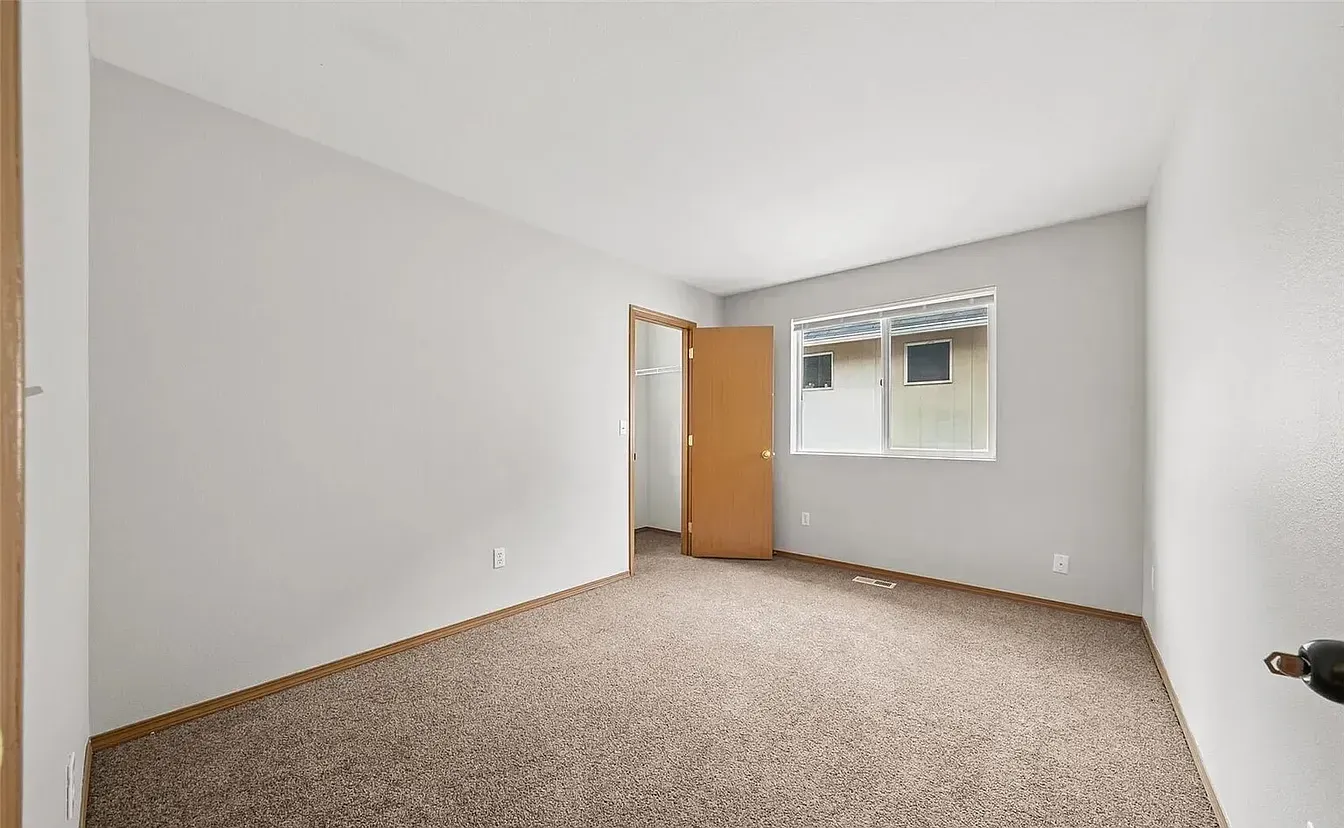 Empty bedroom with beige carpet, light gray walls, window, and closed door.