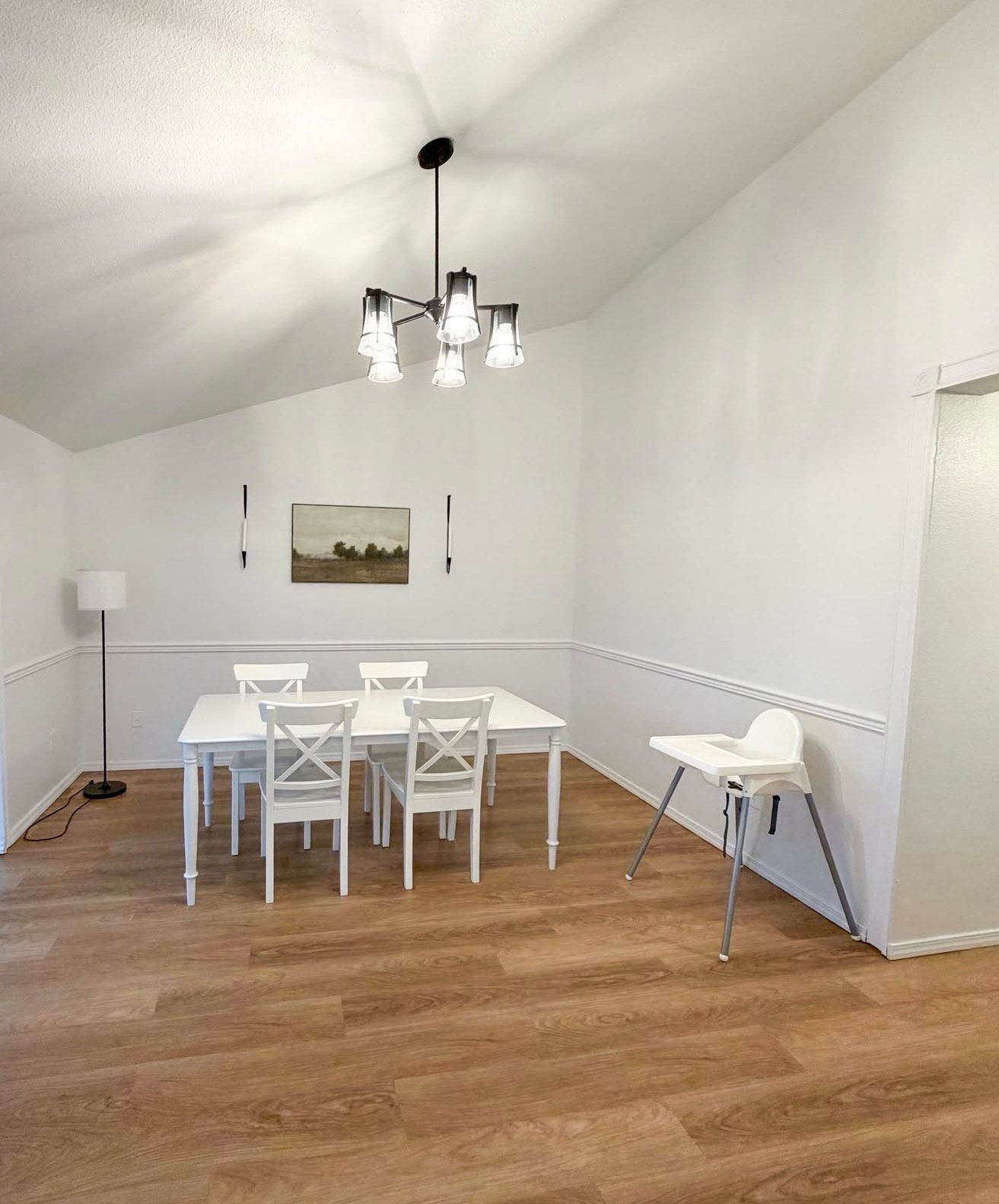 White dining room with a table, chairs, chandelier, and a high chair on wood-look floors.